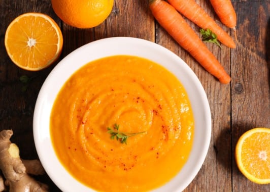 Velouté de courge et carotte au gingembre dans un bol blanc entouré de carottes fraîches, oranges et gingembre